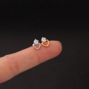 Delicate heart-shaped stainless steel stud earrings with opal stone shown on a fingertip