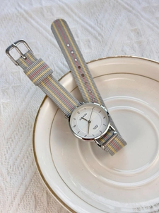 Detail of quartz watch with grey woven fabric strap and silver dial on a porcelain plate