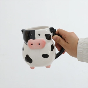 Close-up of hand holding a 3D cow-shaped 20oz ceramic mug with black spots and pink nose