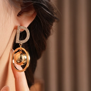 Close-up of gold earrings with rhinestone-studded letter D and spherical pendant on a model