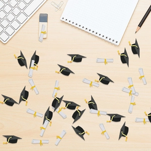 Graduation party confetti with black caps and white scroll shapes on wooden desk