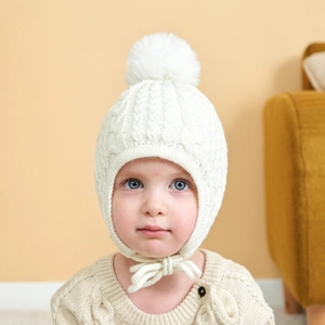 Close-up of white cable knit baby hat featuring plush pompom and warm earflaps with ties