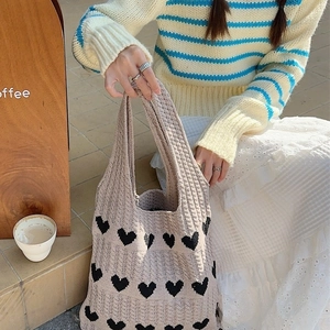Beige knitted tote bag with black heart patterns held by a model in a cozy cafe setting