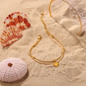 Detail of 18K gold double-layer anklet with heart pendant and beads on beach sand