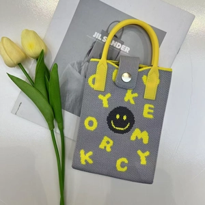Detail of grey knitted phone bag with yellow handle and smile logo beside tulips