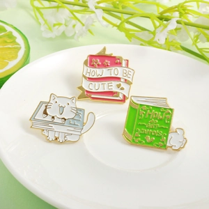 Three enamel pins featuring cute animals on books like a white cat on a blue fish book