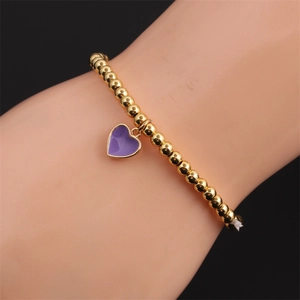 Detailed view of a gold beaded elastic bracelet with a purple enamel heart charm on a wrist