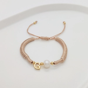 Detail of handmade woven bracelet with pearl and golden letter G charm on white tray