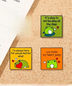 Close-up of colorful cartoon frog enamel pins with inspirational quotes on a cork board