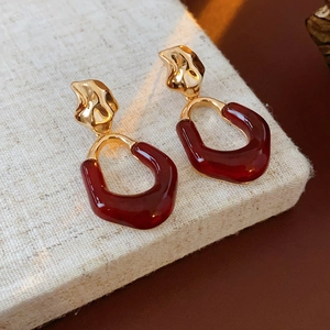 Close-up of textured gold metal earrings with deep red U-shaped resin drops on beige fabric