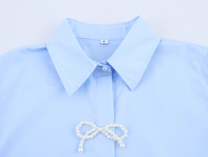 Detail eines hellblauen Hemdkragens mit dekorativer Perle-Schleife und Größenetikett S