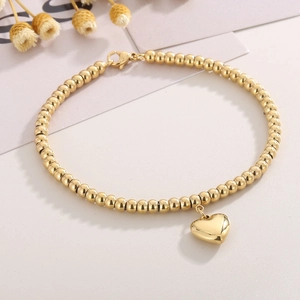 Poliertes goldenes Perlenarmband mit Herzanhänger und Karabinerverschluss auf hellem Grund