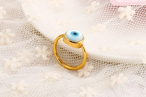 Macro view of gold Evil Eye ring with blue enamel inlay on a white lace background