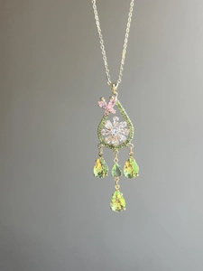 Close-up of gold drop pendant with white flower, pink butterfly and green tassels