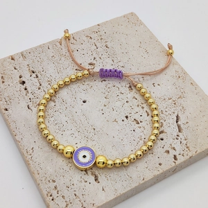 Detail of gold beaded bracelet with purple evil eye charm and adjustable woven cord