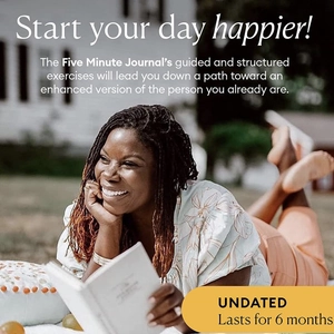 A woman reads the linen Five Minute Journal on grass with a blurred background