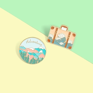 Detailed enamel adventure pins featuring snowy mountains and ocean waves on a suitcase
