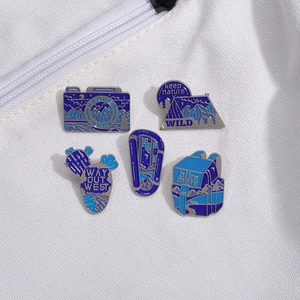 Outdoor adventure enamel pin set featuring camera, tent, and hiking gear on white fabric