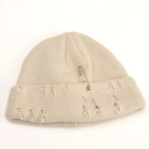 Close-up of beige knit beanie with silver rings, metal pins, and distressed hole details