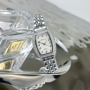Silver tonneau watch with crystal bezel and steel strap displayed next to perfume