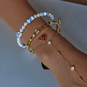 Dettaglio di bracciali dorati sovrapposti con ciondolo a cuore rosso e perle di porcellana
