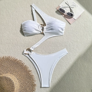 Flat lay of a white one-shoulder cutout swimsuit with gold ring detail on textured background