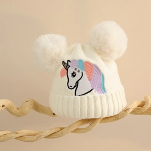 Nahaufnahme der weißen Strickmütze mit Einhorn-Stickerei und flauschigen Doppel-Pompons