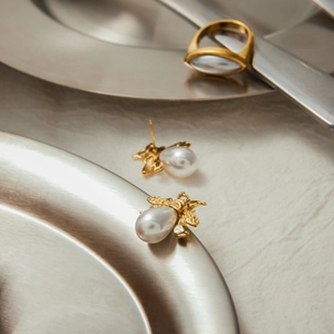 Macro shot of bee-shaped pearl earrings and gold ring on a brushed metal tray