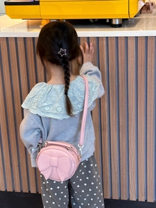Pink round crossbody bag for children with large bow detail shown in a cafe setting