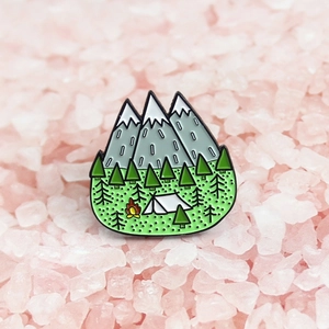 Macro view of camping enamel pin with snowy mountains, pine trees, and a campfire detail