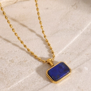 Close-up of golden beaded chain necklace with rectangular blue gemstone pendant on stone