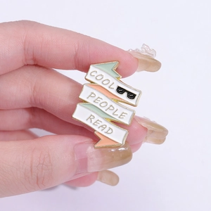 Hand holding a Cool People Read enamel pin with book design and gold metal frame