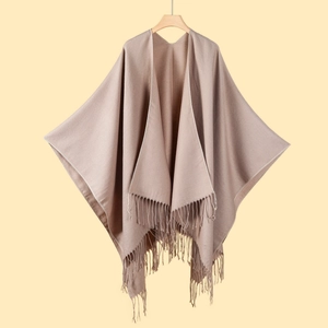 Light brown knit shawl cape with fringe tassels on wooden hanger against warm background