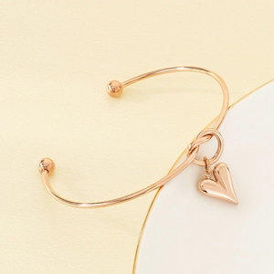 Detail of rose gold heart charm open bangle with bead ends on a beige and white background