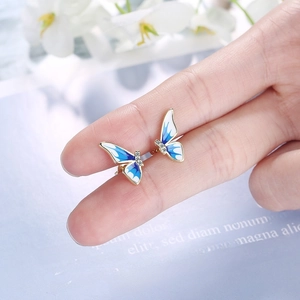 Golden butterfly stud earrings with blue enamel wings and crystals held on a finger