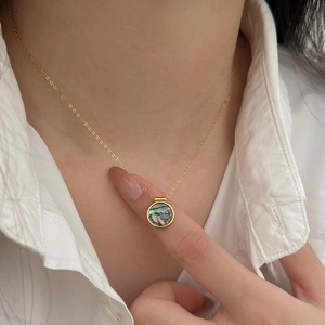 Close-up of a round abalone shell pendant on a gold chain necklace worn over a white shirt