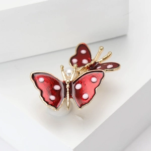 Red enamel twin butterfly brooch with golden edges and a central pearl on a white base