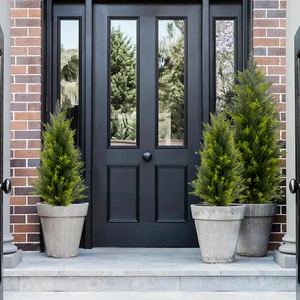 Three potted artificial cypress trees in grey planters set against a red brick wall