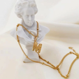 Macro detail of a gold textured letter M pendant on a beaded stainless steel chain