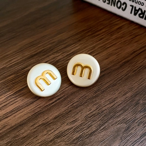 Macro shot of circular cream enamel earrings with golden letter M logo on wood surface