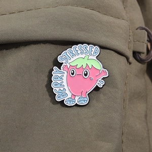 Close-up of pink strawberry enamel pin on green canvas bag showing Berry Stressed text