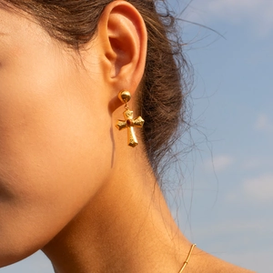 Close-up of gold cross earring with hammered texture worn by a model against a blue sky