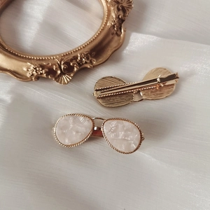 Elegant gold sunglasses-shaped hair clip with white shell inlay on soft fabric background