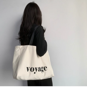 Rear view of a model carrying a large cream canvas tote bag with black voyage lettering