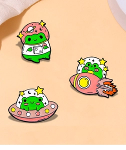 Detailed view of three cartoon space frog enamel pins on a light orange background