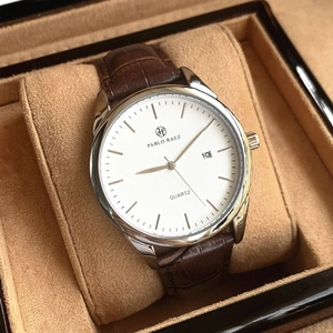 Detail view of quartz watch with silver case, white dial, and brown leather strap in box