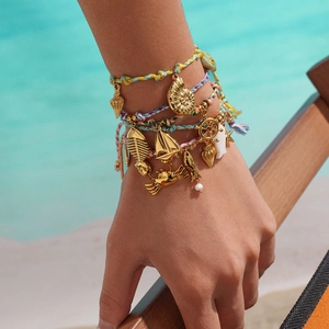 Detail of colorful woven ocean bracelet with gold charms on a model's wrist at the beach