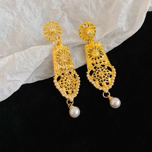 Detail of long hollow patterned gold earrings with white pearls on a dark velvet texture