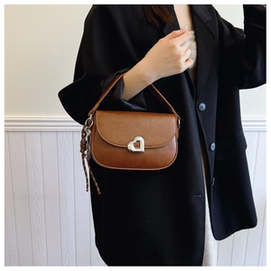 Detail of a brown leather saddle bag with a silver heart-shaped clasp and woven knot tassel