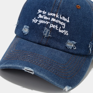 Close-up of distressed denim baseball cap featuring white embroidered quote and torn details